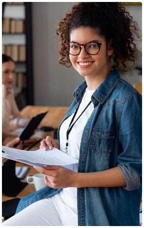 About Us 2 Young woman with curly hair and glasses, wearing a denim jacket, smiles while holding papers in a bright office. A person works at a laptop in the background.